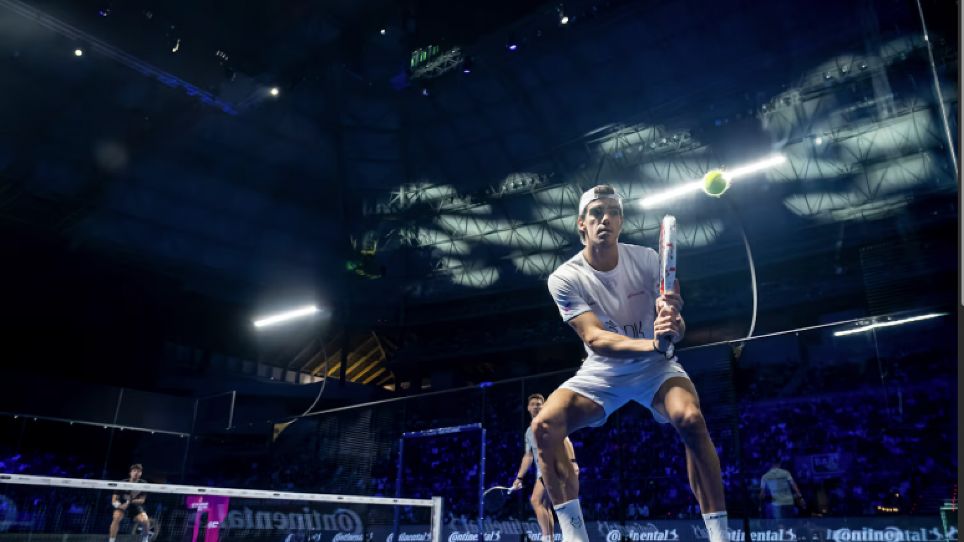 Action Moment at Padel A professional padel player prepares for a fast volley during a high-intensity match inside a large indoor arena with dramatic lighting and a full crowd.